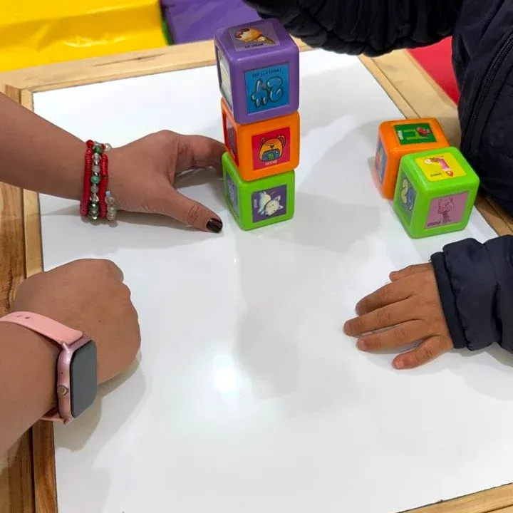 Un niño y la maestra apilan cubos de colores con dibujos sobre una mesa blanca.