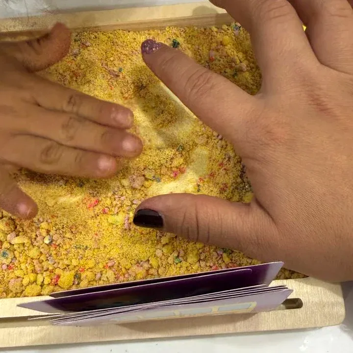Las manos de la maestra y de un niño trazan juntas en una bandeja de arena de colores.