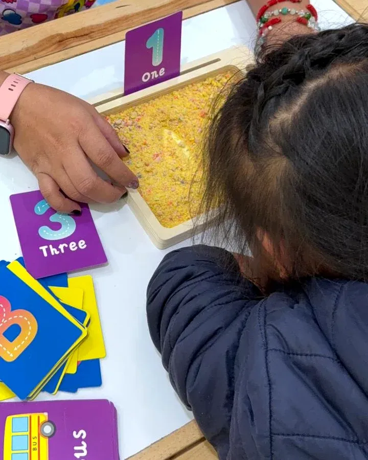 Una niña, de espaldas, traza en la bandeja de arena junto a las tarjetas de los números 1 y 3.