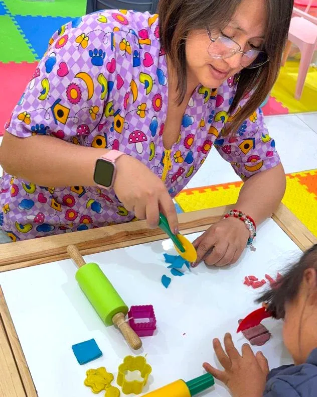 La maestra corta plastilina con un molde mientras una niña amasa la suya con rodillos y cortadores en la mesa.