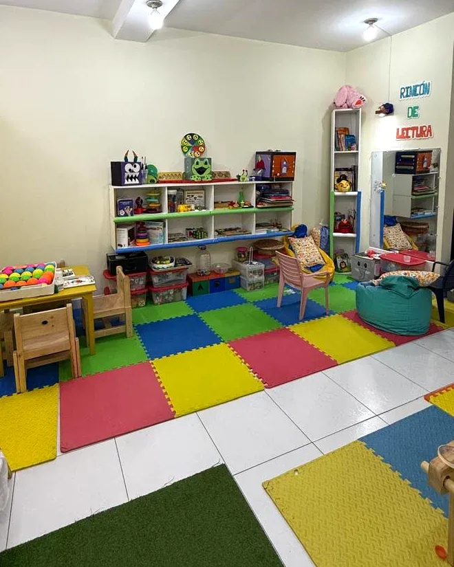Rincón de lectura con libreros, cojines, un puf turquesa y una mesa amarilla con material para contar.