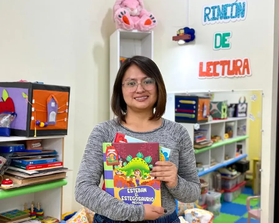 Michelle Palma sonríe en el rincón de lectura de ParvuArt, con suéter gris y lentes, sosteniendo varios libros infantiles; detrás, estantes con juegos y un conejo de peluche rosa.