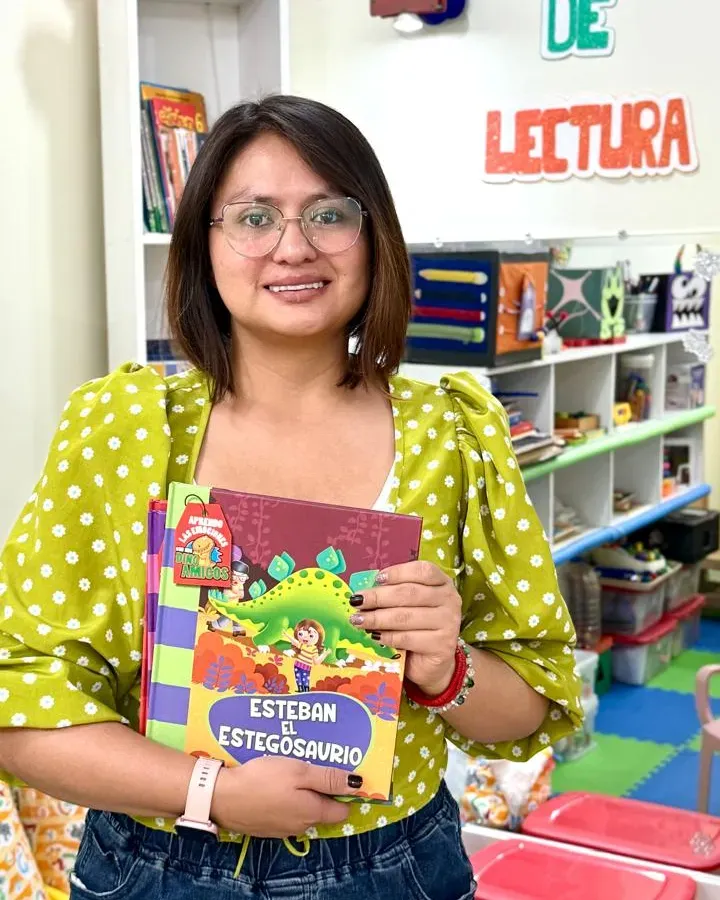 Michelle Palma, con blusa verde de lunares y lentes, sonríe sosteniendo un libro infantil con un dinosaurio verde en la portada, en el rincón de lectura.